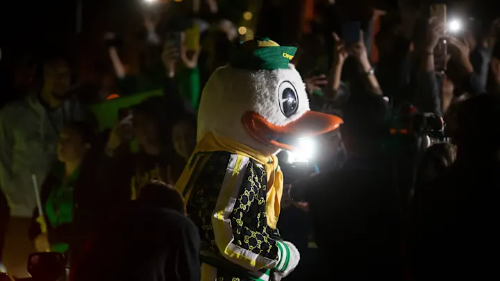 The Oregon Duck mascot surrounded by fans with phone flashlights at a night College GameDay